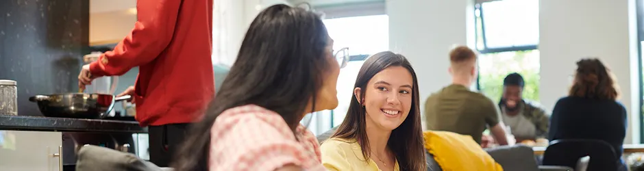 Female college students talking sitting on couch. Male student cooking behind them